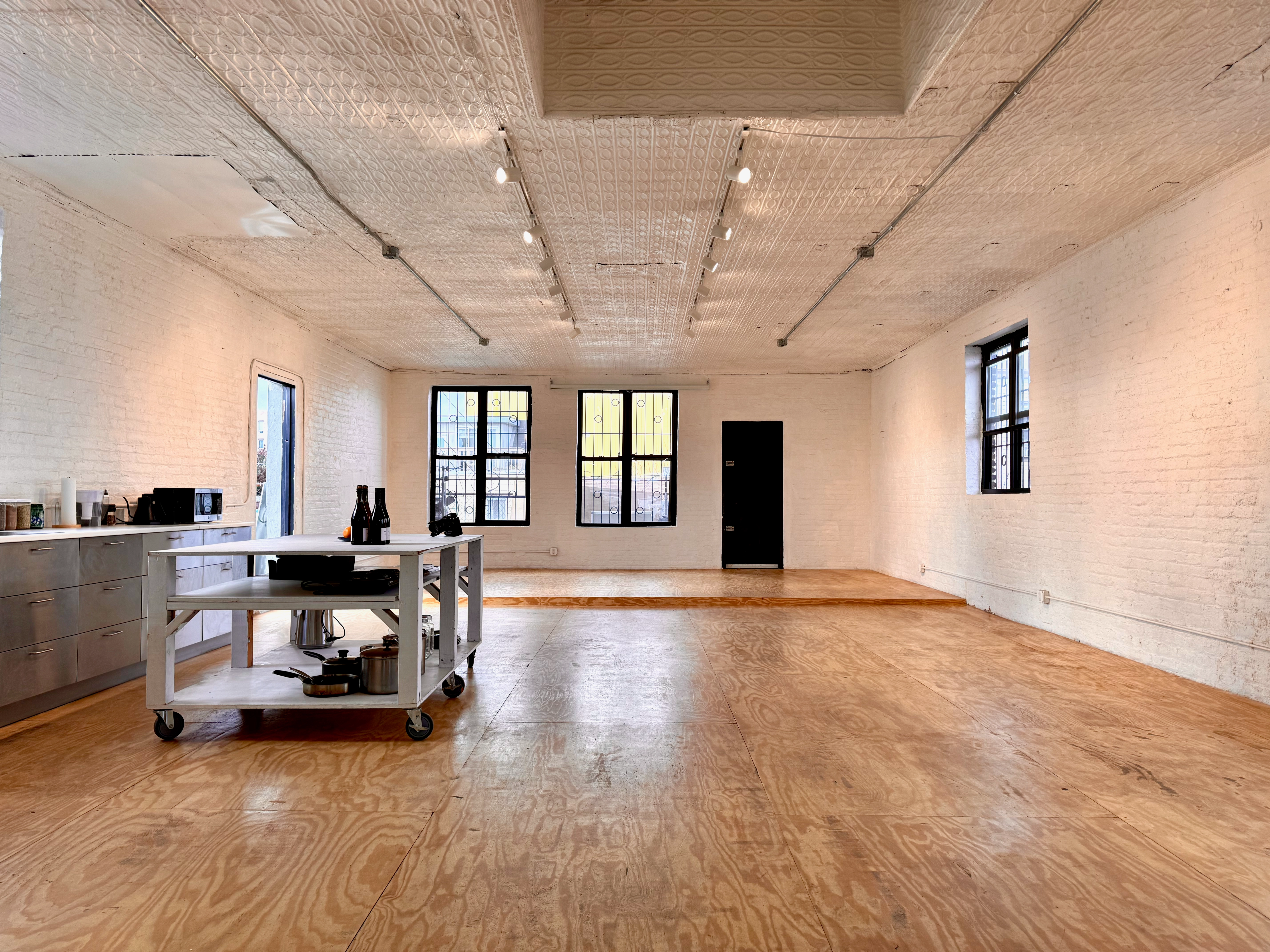 A wide-angle photo of the sunny telos.haus kitchen, including a large white island. In the background, a stage. The plywood floors gleam in the light, and various supplies and cookware are visible in the island shelves.