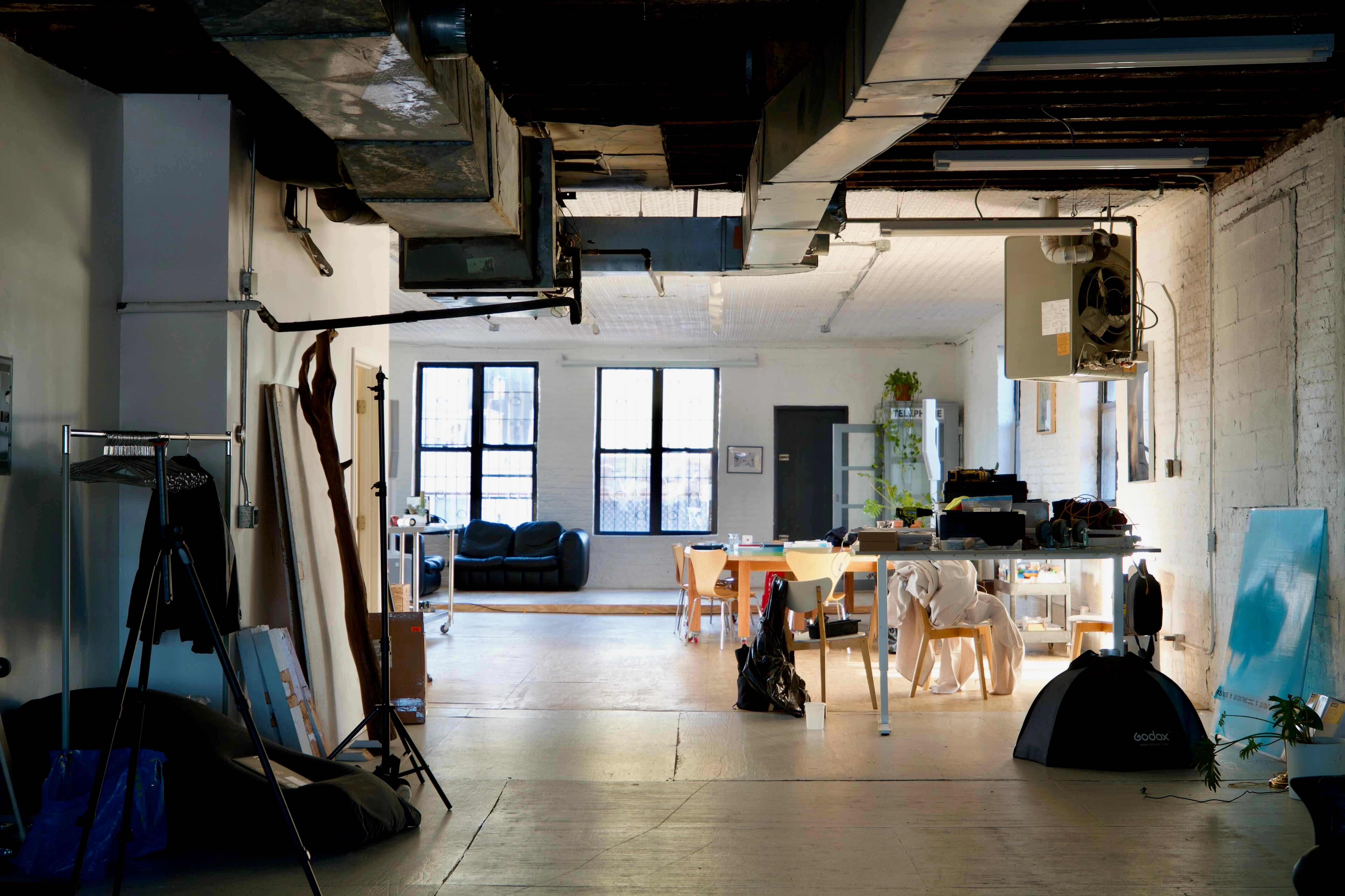 A wide-angle photo of the sunny telos.haus kitchen, including a large white island. In the background, a stage. The plywood floors gleam in the light, and various supplies and cookware are visible in the island shelves.